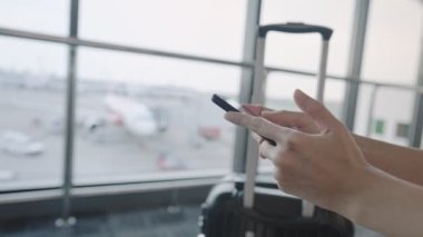 Akıllı telefon kullanan eller uçağa binmeden önce havaalanı terminalinde bekler, seyahat bileti rezervasyonu, seyahat valizi, Airlines Aerobridge ve arka planda uçaklar.