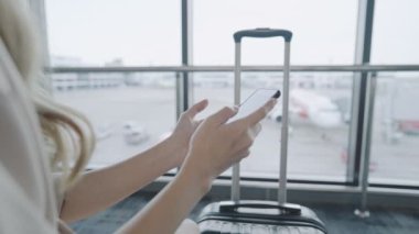 Zarif Sarışın Kadın elleri havaalanı terminalinde cep telefonuyla oturur havaalanı terminalinde cep telefonu kullanır, havaalanı terminalinde cep telefonu kullanır, seyahat valiziyle havayolu seyahati yapar, bilet ayırtır.