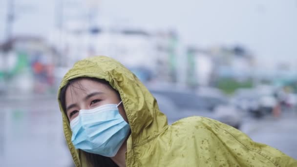 若いかなりアジアの女の子は 道路側に立って黄色のレインコートを保護顔マスクを着用し 車の背景に都市部の通りを止め 汚染された都市 雨季の気候で安全に滞在 — ストック動画