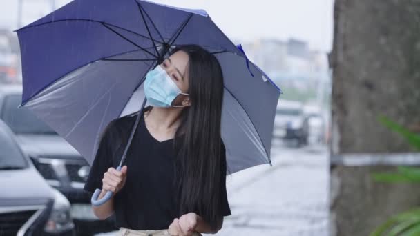 若いアジアの女性は 道路脇の散歩に立って青い傘を保持保護顔マスクを着用 雨を注ぐ 雨を注ぐ 病気になるリスク ハード雨を注ぐ 手好奇心に達する — ストック動画