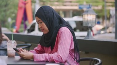 Çekici Asyalı Müslüman tek başına kahve masasında oturuyor cep telefonu uygulaması kullanıyor, çevrimiçi mesajlaşma alıyor, sosyal medya kuruyor, telefon ekranında okuyor, tesettüre dayalı geleneksel giysiler giyiyor, modern teknoloji ve yaşam eğlencesi sunuyor. 