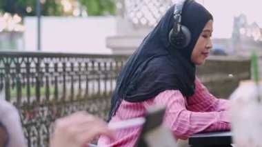 Genç Güneydoğu Asyalı Müslüman kız tesettüre giriyor, kulaklık takıyor kahve dükkanında boş zaman geçiriyor, öğleden sonra dinleniyor, modern teknoloji cihazları, taşınabilir uygulama kullanıcısı, yoğun günlerde kahve molası, modern teknoloji ve yaşam eğlencesi