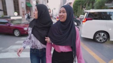 Çekici genç Asyalı kadın Müslümanlar arkalarda hareket halindeki arabalarla yaya geçidinde yürürken kollarını tutuyorlar. Kadınlık toplumu, birlikte modern şehri ziyaret eden heyecanlı turistler, gezici yürüyüşler, modern insanlar iyi ilişkilerden keyif alıyorlar.