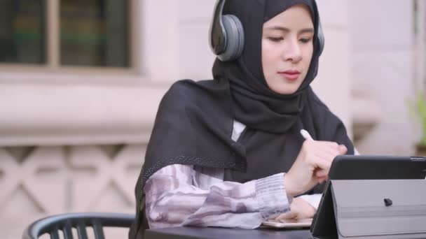 ポータブルミニタブレットとスマートペン屋外に座って作業ヘッドセットを身に着けているアジアのイスラム教徒の女性 近代的な技術 タブレットを見てスマートライフ 遠隔作業場所 ワイヤレスインターネット接続 現代の女性のイスラム教徒と生活ライフスタイル — ストック動画