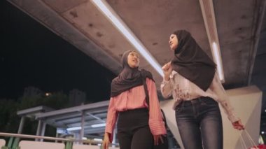 İki mutlu Güneydoğu Asyalı Müslüman kız neşeli bir şekilde modern bir şehir köprüsünün altına doğru yürüyorlar, kültürel birlik dostluğu, mutlu ve gülümseyerek haftasonu boyunca olumlu bir konuşma yapıyorlar, modern insanlar iyi bir ilişkiden zevk alıyorlar.