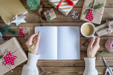 Masaüstü planlayıcısı. Bir deftere bir kadın eli yazıp Noel hediyesini paketliyor. Dekor unsurlarıyla ahşap arka planda sunumlar, üst görünüm. Yeni yıl paketleme kavramı, üst görünüm.