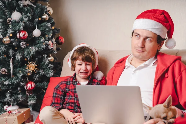 Family in Santa hat, father and crying child boy with laptop sitting on a couch in the living room with dog.