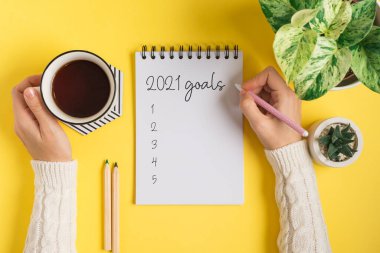 2021 gol mesajı yazan boş not defterli kadın elleri. Sarı çalışma masasının arka planında bir fincan çay ve bitkilerle yatıyordu. Üst görünüm modeli 