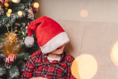 Kırmızı şapkalı şirin çocuk Noel Baba 'yı evde Noel ağacıyla oturma odasında bekliyor.