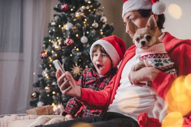 Noel tatilinde elinde cep telefonuyla evde Noel ağacı olan oturma odasındaki koltukta oturan çocuklu ve yavru köpekli baba ve Noel Baba..