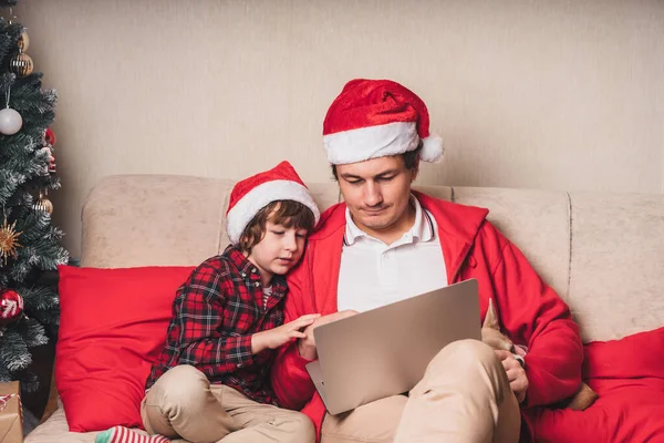 Πατέρας με παιδί και κουτάβι σε καπέλα Σάντα έχοντας μια βιντεοκλήση την ημέρα των Χριστουγέννων στο φορητό υπολογιστή, κάθεται σε έναν καναπέ στο σαλόνι με χριστουγεννιάτικο δέντρο στο σπίτι. — Φωτογραφία Αρχείου