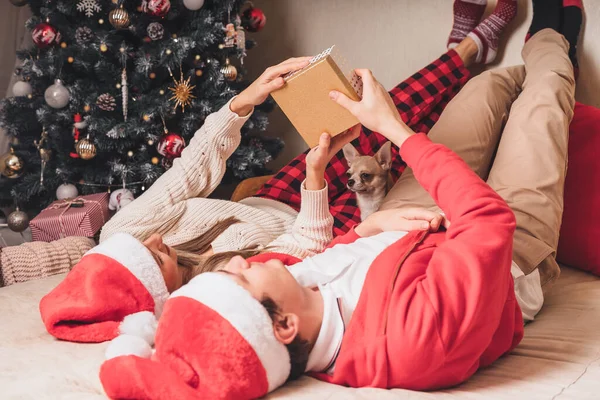 Romantic couple exchanging gifts near decorated christmas tree. Man giving christmas present surprise to smiling woman in sweater while relaxing on couch. Boyfriend and girlfriend. — Stock Photo, Image