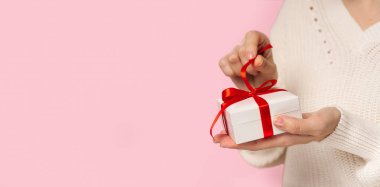 Güzellik Kadını el ele tutuşup pembe arka planda kırmızı fiyonklu hediye kutusunu açıyor. Yakın plan. Pastel renkler, metin için alanı kopyalayın. Sevgililer günü hediyesi. Noel bayrağı, doğum günü, doğum günü konsepti