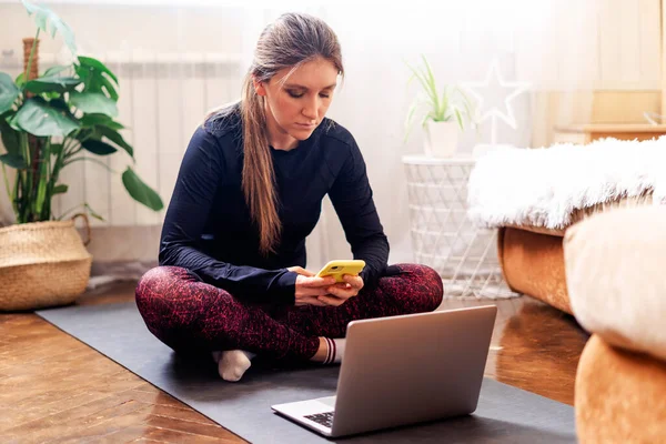 Спортивная женщина в спортивной одежде работает и использует ноутбук дома в гостиной, сидит на полу с гантелями на бирюзовом коврике и слушает музыку. Концепция спорта и отдыха — стоковое фото