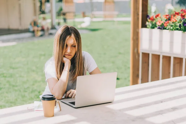 Vacker kvinna som jobbar på notebook sätter sig på bänken utanför och dricker kaffe. Använda datorer och nätbutiker. Glad tjej med ljust hår. Distansutbildning på nätet — Stockfoto