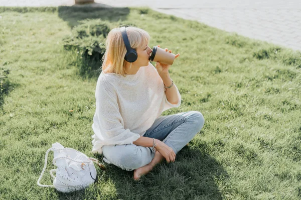 Junge Frau mit Kopfhörern sitzt draußen im Park auf dem Gras — Stockfoto