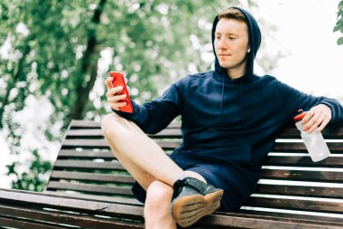 Kapüşonlu genç adam antrenmandan sonra bankta oturup cep telefonu kullanarak rahatlıyor. Yazın güneşli yeşil parkta egzersiz yaparken su şişesini tutmak.
