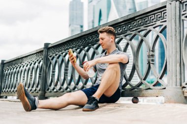 Yolda oturan yorgun genç adam, cep telefonu kullanıyor ve spor eğitiminden sonra dinleniyor. Şehir caddesinde spor yaparken su şişesini tutmak, yazın gökyüzü bulutlu.