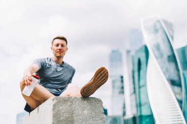Merdivenlerde oturup spor antrenmanlarından sonra dinlenmekten yorgun düşmüş genç adam. Yaz kenti şehir sokağında spor yaparken su şişesini tutmak, bulutlu bir gökyüzü..