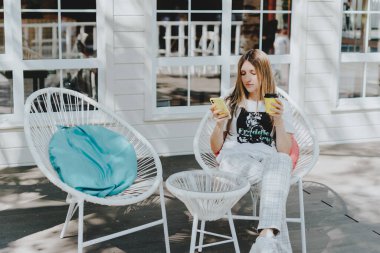 Genç bir kadın cep telefonuyla görüntülü konuşma yapıyor ve kahve içiyor. Yazlık kafede sandalyede oturan mutlu kız. Cep telefonunda teknoloji ve çevrimiçi mesaj kullanılıyor