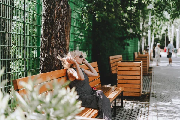 Frau sitzt auf Holzbank im Sommerpark mit grünen Bäumen und sonnigem Hintergrund mit Gras und Pflanzen. Telefonieren. — Stockfoto