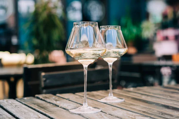 A wooden table in a restaurant with white wine. Wine glasses served for a party in a bar or a restaurant on terrace at a sunny day. Blurred background with plants. Still life scene. — Stock Photo, Image