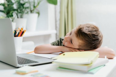 Uzaktan öğrenme çevrimiçi eğitim. Kafkasyalı çocuk laptopta ders çalışıyor, uyumaktan ve not defterinde uyumaktan yoruluyor, okul ödevlerini yapıyor. Elinde defter ve eğitim kitabı olan bir çocuk. Okula geri dön.