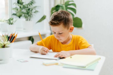 Uzaktan öğrenme çevrimiçi eğitim. Kafkasyalı gülümseyen çocuk evde kitap okuyor, not defterine yazıyor ve ödev yapıyor. Çocuğun elinde defterle masada oturduğunu düşün. Okula geri dön.