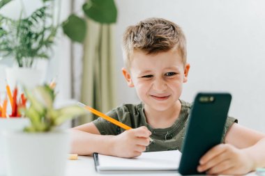 Okula dönelim. Uzaktan öğrenme çevrimiçi eğitim. Kafkasyalı gülümseyen çocuk cep telefonuyla evde ders çalışıyor ve okul ödevi yapıyor. Somurtkan çocuk cep telefonu ve kalemle masada oturuyor.