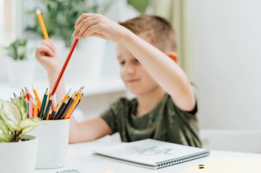 Uzaktan öğrenme çevrimiçi eğitim. Kafkasyalı gülümseyen çocuk evde kitap okuyor, not defteri çiziyor ve okul ödevi yapıyor. Çocuğun elinde defterle masada oturduğunu düşün. Okula geri dön.