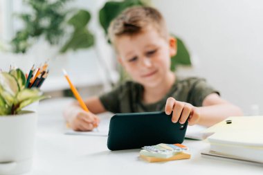 Okula dönelim. Uzaktan öğrenme çevrimiçi eğitim. Kafkasyalı gülümseyen çocuk evde cep telefonuyla ders çalışıyor ve okul ödevi yapıyor. Cep telefonu, kalem ve eğitim kitabı olan bir çocuk.