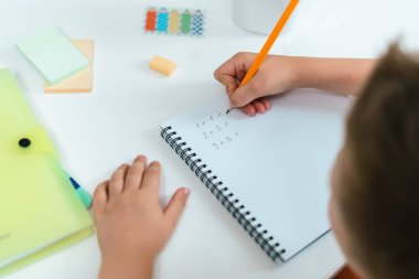 Uzaktan öğrenme çevrimiçi eğitim. Çocuk evde kitapla çalışıyor, not defterine yazıyor ve okul ödevi yapıyor. Çocuk elinde not defteriyle masada oturuyor. Matematiksel problemleri çözmek. Okula geri dön.