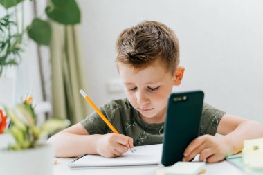 Okula dönelim. Uzaktan öğrenme çevrimiçi eğitim. Kafkasyalı gülümseyen çocuk evde cep telefonuyla ders çalışıyor ve okul ödevi yapıyor. Cep telefonu, kalem ve eğitim kitabı olan bir çocuk.