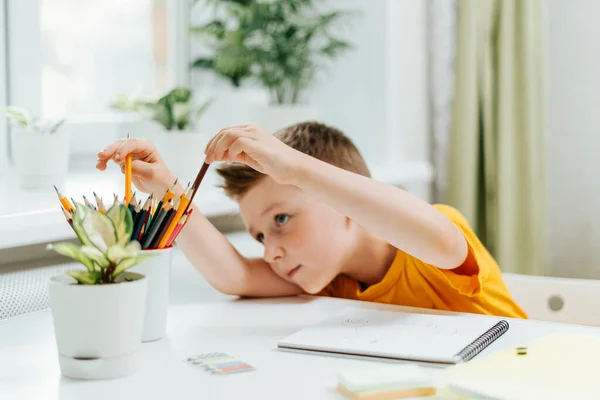 Enseignement à distance en ligne. Garçon sourire caucasien étudiant à la maison avec le livre, dessiner dans le bloc-notes et faire des devoirs scolaires. Pensant enfant assis à table avec ordinateur portable. Retour à l'école — Photo