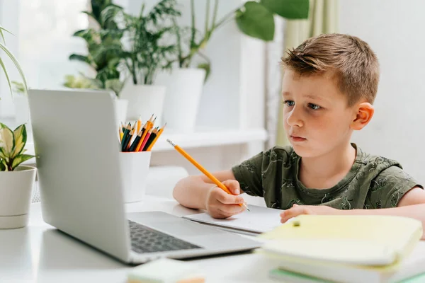 Enseignement à distance en ligne. Blanc sourire garçon enfant étudiant à la maison avec ordinateur portable et faire des devoirs scolaires. Pensant à l'emplacement des enfants avec cahier, crayons et livres de formation. Retour à l'école — Photo