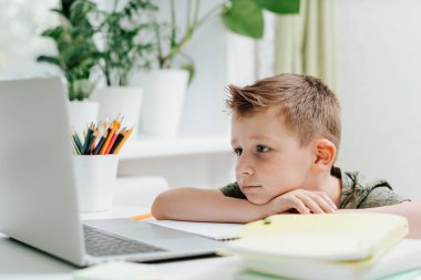 Uzaktan öğrenme çevrimiçi eğitim. Kafkasyalı gülümseyen çocuk evde dizüstü bilgisayarla ders çalışıyor ve okul ödevi yapıyor. Düşünen çocuk elinde defter, kalem ve eğitim kitaplarıyla oturuyor. Okula geri dön.