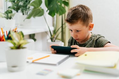Okula dönelim. Uzaktan öğrenme çevrimiçi eğitim. Kafkasyalı gülümseyen çocuk evde cep telefonuyla ders çalışıyor ve okul ödevi yapıyor. Cep telefonu, kalem ve eğitim kitabı olan bir çocuk.