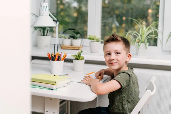 Distansutbildning på nätet. Vit leende pojke som studerar hemma med bok, skriver anteckningsblock och gör läxor. Leende barn sittandes vid bordet med anteckningsbok. Tillbaka till skolan — Stockfoto