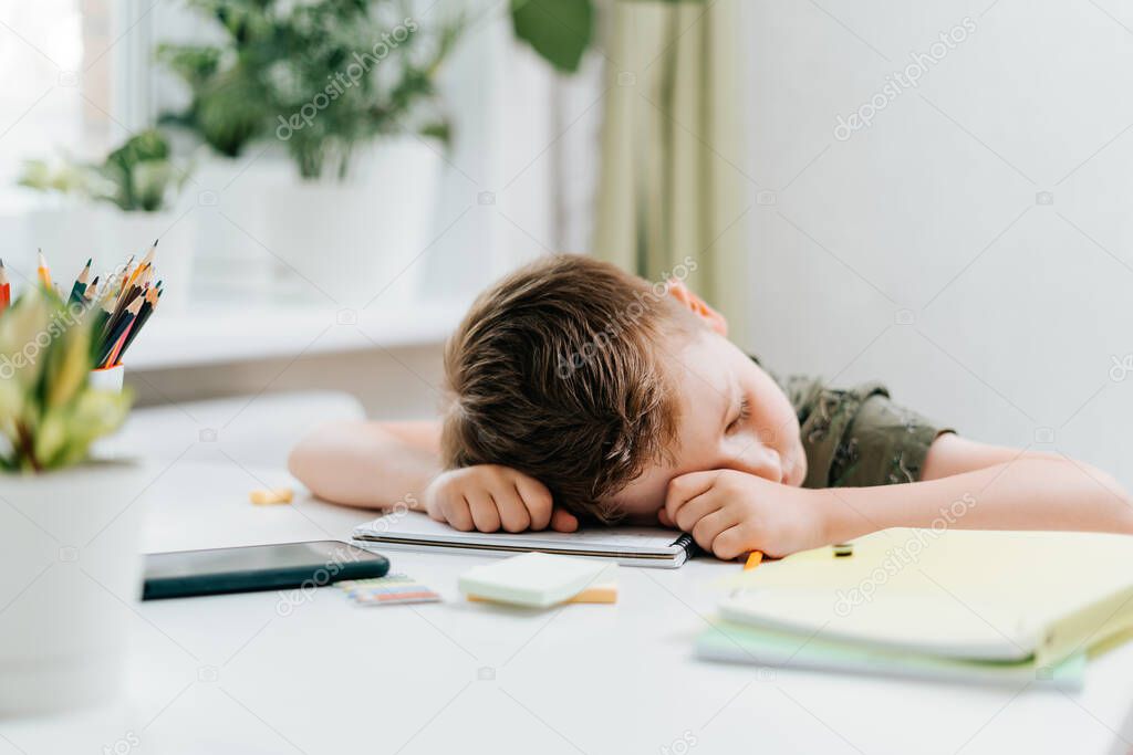 Distance learning online education. Caucasian kid boy study at home, tired asleep, resting his head on notepad, exhausting homework. Child siting with notebook and training book. Back to school