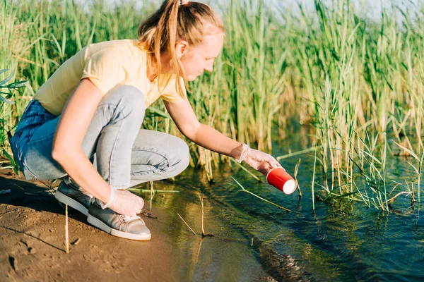 Jovem voluntária satisfeita com a coleta de lixo, garrafas de plástico e xícaras de café, limpe a praia com um lago. Mulher recolhendo lixo no saco. Conceito de poluição ecológica ambiental — Fotografia de Stock