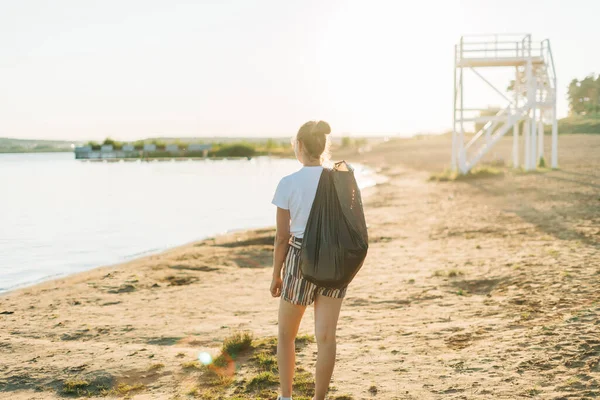 Joven voluntaria con bolsa y guantes recogiendo basura, botellas de plástico, limpiando la playa con un mar. Mujer recogiendo basura. Ecología ambiental concepto de contaminación. Día de la Tierra —  Fotos de Stock