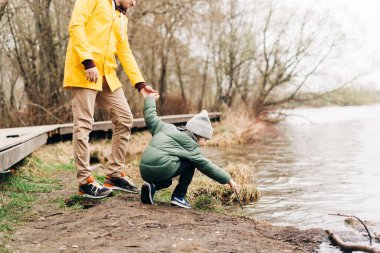 Baba ve oğul gölün kıyısında durup suyla oynuyorlar. Sonbahar parkında oyun oynayan ve dışarıda eğlenen çocuklu mutlu bir aile.