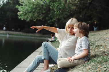 Anne ve oğlu parkta nehir kıyısında oturuyorlar. Çocuk ve annesi çimlerin üzerinde gölet kıyısında dinleniyorlar. Kadın elini göle doğrultuyor. Aile dışı aktiviteler su kenarında