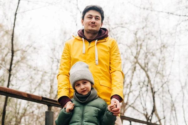 黄色のレインコートを着た父と森で楽しんでいる息子。子供の男の子が遊んでいて、秋の公園の背景で屋外で楽しんでいる幸せな家族 — ストック写真
