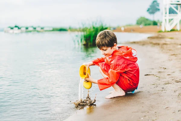 Niño en un impermeable rojo vierte agua de bota de goma amarilla en el lago. Niño jugando con agua en el estanque. Niño divirtiéndose al aire libre cerca de la orilla del río —  Fotos de Stock