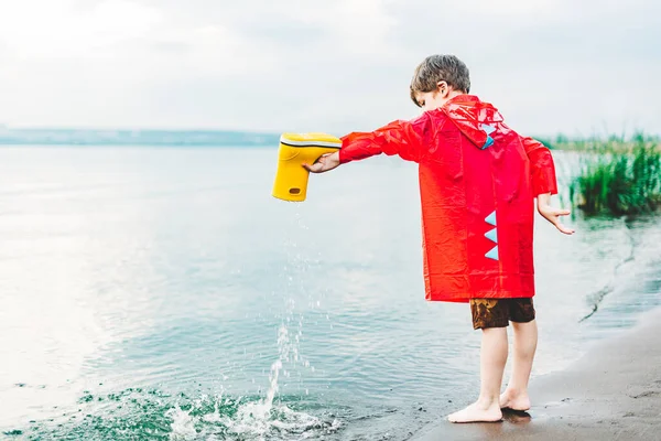 Um rapaz de gabardina vermelha derrama água da bota de borracha amarela para o lago. Criança brincando com água na lagoa. Criança se divertindo ao ar livre perto da ribeira — Fotografia de Stock