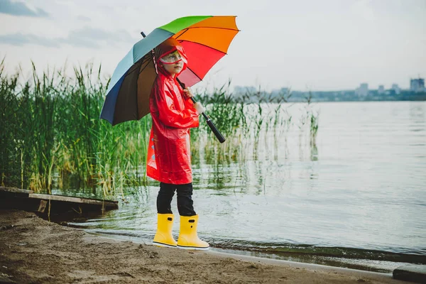 Ragazzo in impermeabile rosso e stivali di gomma gialla si trova sulla riva del fiume e tiene l'ombrello arcobaleno. Ragazzo della scuola fermo vicino al lago autunnale. Bambino che indossa vestiti impermeabili a riva — Foto Stock