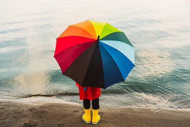 Kırmızı yağmurluk ve sarı lastik çizmeli çocuk kumsalda duran gökkuşağı şemsiyesini tutuyor. Renkli şemsiyeli çocuk denize bakıyor. Su geçirmez kırmızı ceketli okul çocuğu elinde çok renkli şemsiye tutuyor.