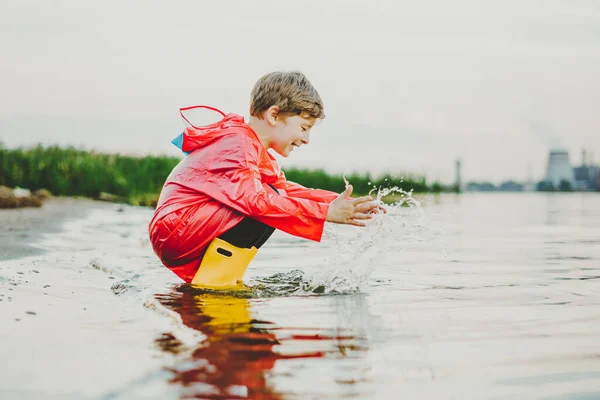 Niño con un impermeable rojo y botas de goma amarillas jugando con agua en la playa. Niño de la escuela con un abrigo impermeable tocando agua en el mar. Niño divirtiéndose con olas en la orilla —  Fotos de Stock
