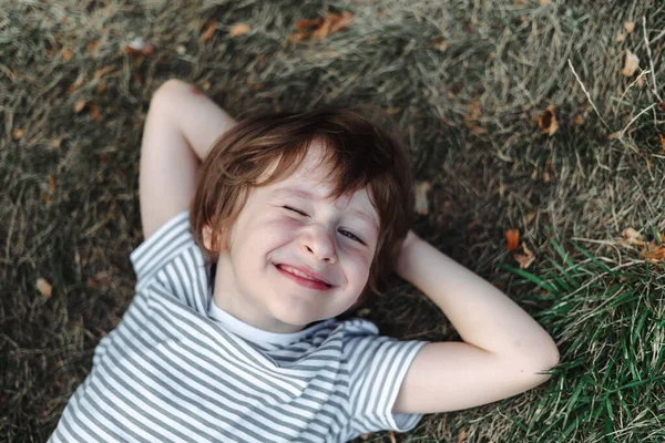 El chico acostado boca arriba en el césped en el parque. Chico relajándose en el césped verde. Niño relajándose afuera, descansando su cabeza en el prado del jardín —  Fotos de Stock